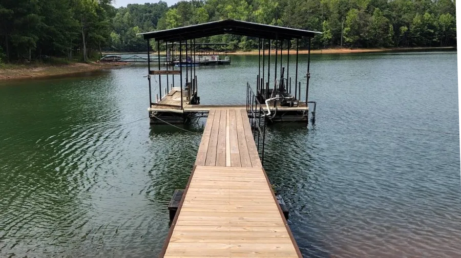 A long wooden dock extends over green water towards two covered boat slips and a tree-lined shore.
